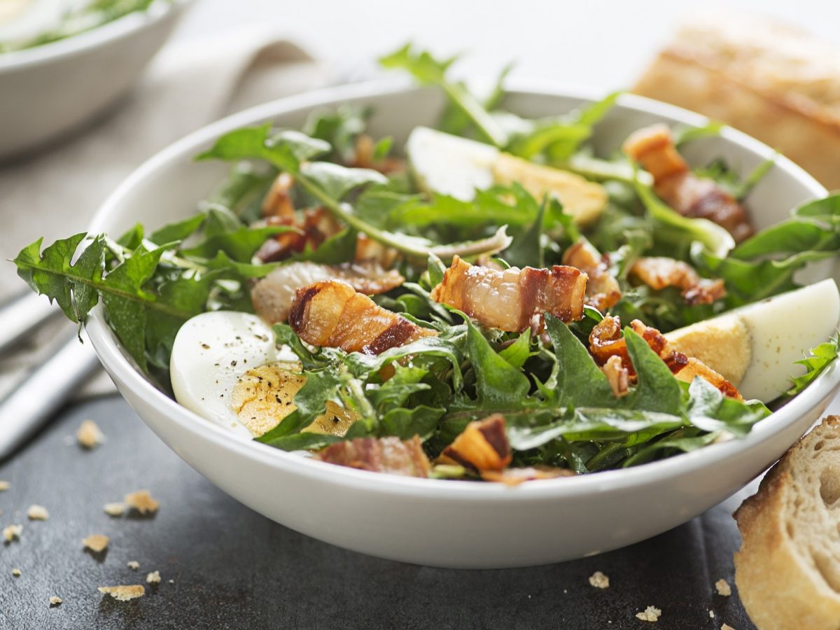 Draufsicht, schräg: eine weiße Schüssel mit Löwenzahn-Salat mit Ei und knusprigen Speck auf dunklem Untergrund. Daneben liegen ein Besteck und Weißbrotscheiben.