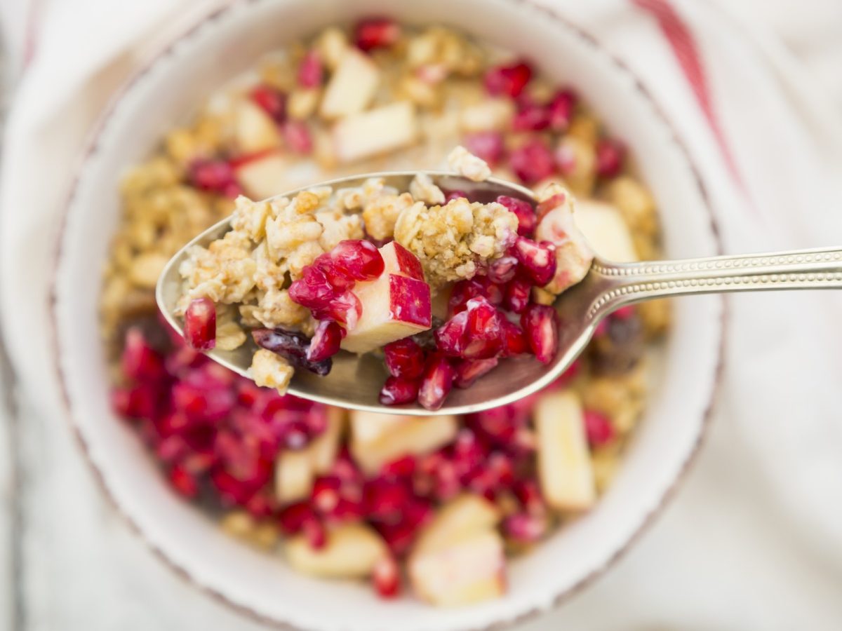 Draufsicht: Eine Schüssel Nuss-Granola mit Granatapfel. Darüber ist ein Löffel mit dem gleichen Granola.