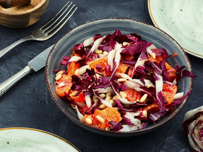 Draufsicht: Auf einem dunklen Untergrund steht eine Schüssel Radicchio-Orangen-Salat. Daneben liegen Messer, gabel, ein Radicchio-Strunk sowie zwei Teller, von denen nur die Ränder zu sehen sind.