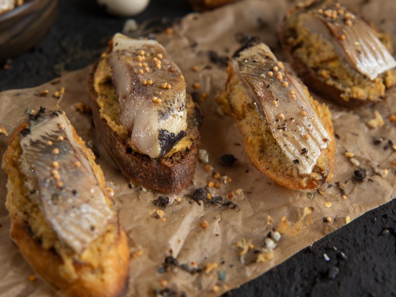 Draufsicht schräg: Ein Backpapier mit Fingerfood. Es handelt sich um mehrere Bruschetta mit Matjes-Hering und eingelegter Senfsaat.