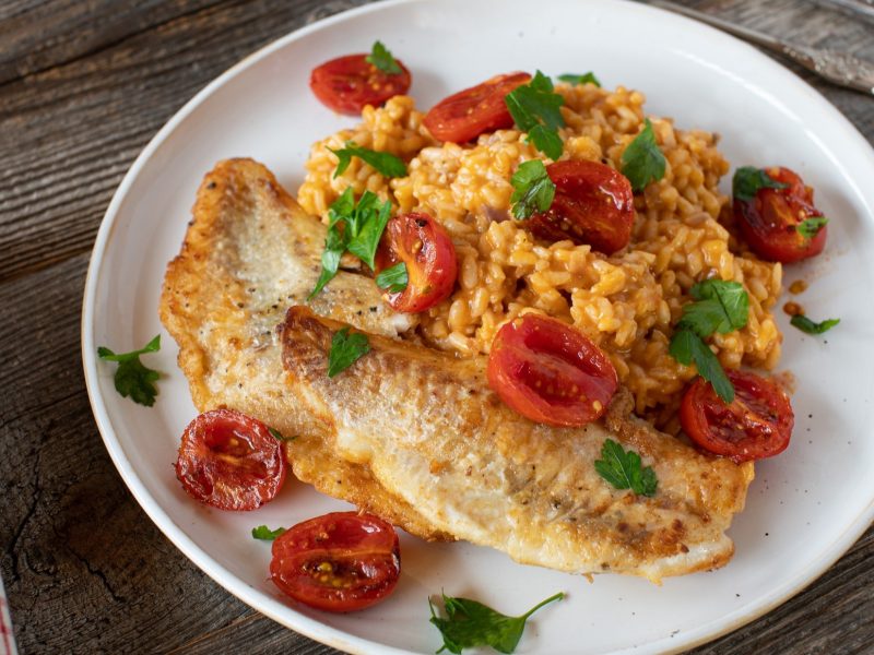 Draufsicht: Auf einem rustikalen Untergrund aus Holz steht ein weißer Teller mit einem Tomaten-Risotto mit Kabeljau. Daneben liegen Besteck und ein kariertes Küchenhandtuch.