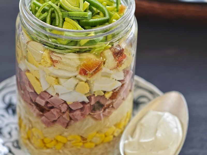 Eiersalat mit Sellerie und Kochschinken im Glas geschichtet auf einem kleinen Teller mit dunklem Untergrund und Löffel.