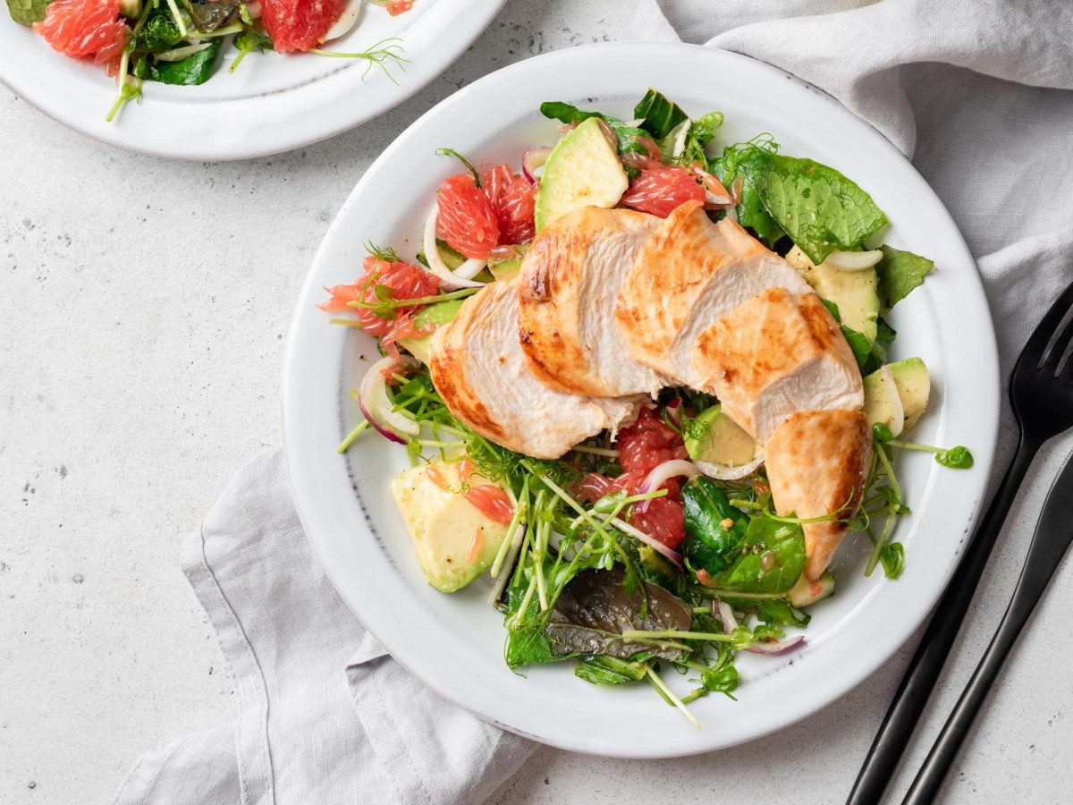 Draufsicht: Ein runder weißer Teller mit einem Hähnchensalat mit Spinat, Avocado und Grapefruit. Daneben liegen Messer und Gabel. Ein weiterer Teller ist teilweise zu sehen.