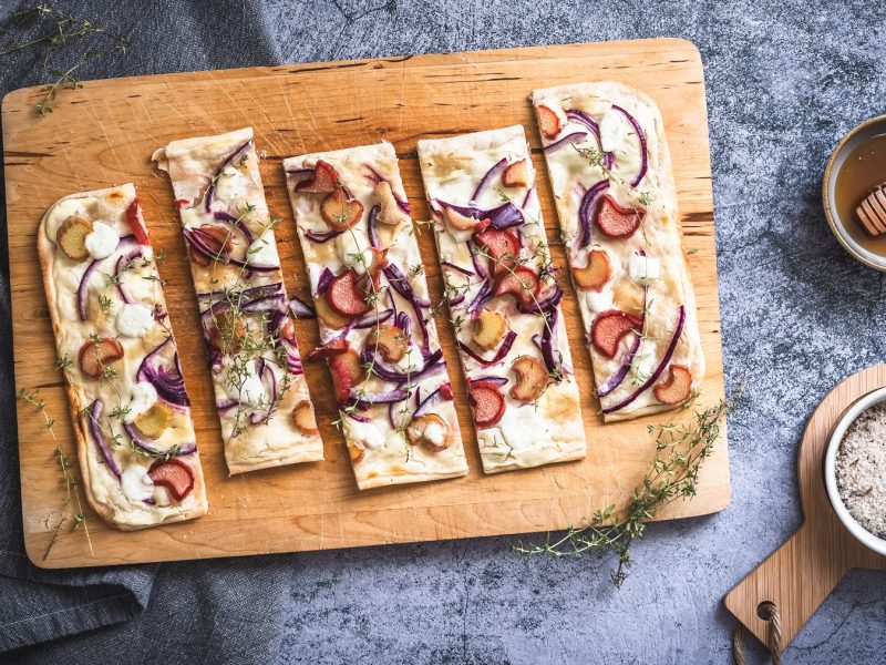 Flammkuchen mit Rhabarber, roten Zwiebeln, Thymian und Ziegenfrischkäse auf einem Holbrett. Daneben eine Schale mit Honig und eine Schale mit Meersalz.