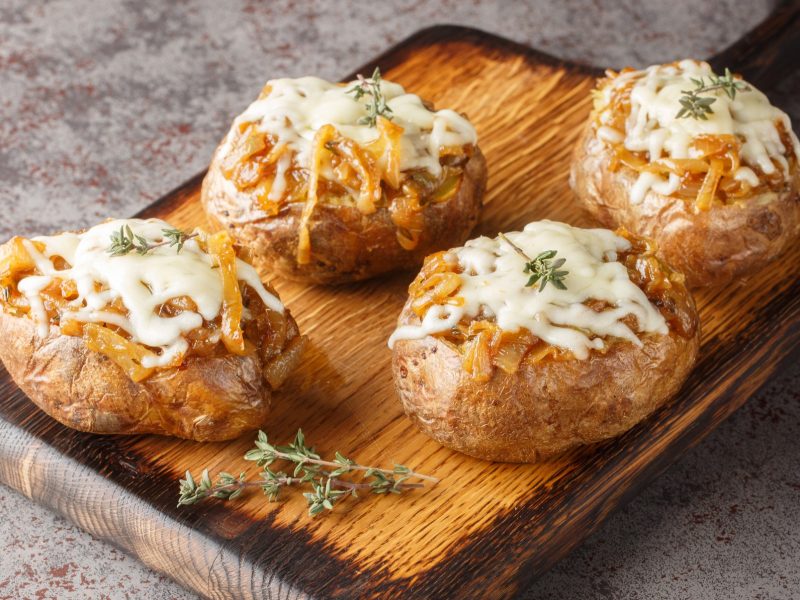 Vier French Onion Potatoes auf einem Holzbrett mit Thymianzweig und grauem Tisch. Alles von schräg oben fotografiert.