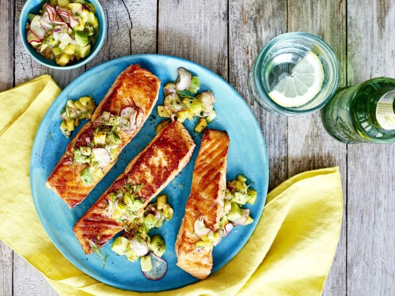 Gewürzlachs mit Radieschen-Avocado-Salsa auf einem blauen Teller auf einem gelben Geschirrtuch. Der Hintergrund ist hölzern. Rechts auf dem Bild eine Wasserflasche und ein Glas Wasser, links auf dem Bild eine kleine Schüssel mit Radieschen-Avocado-Salsa.