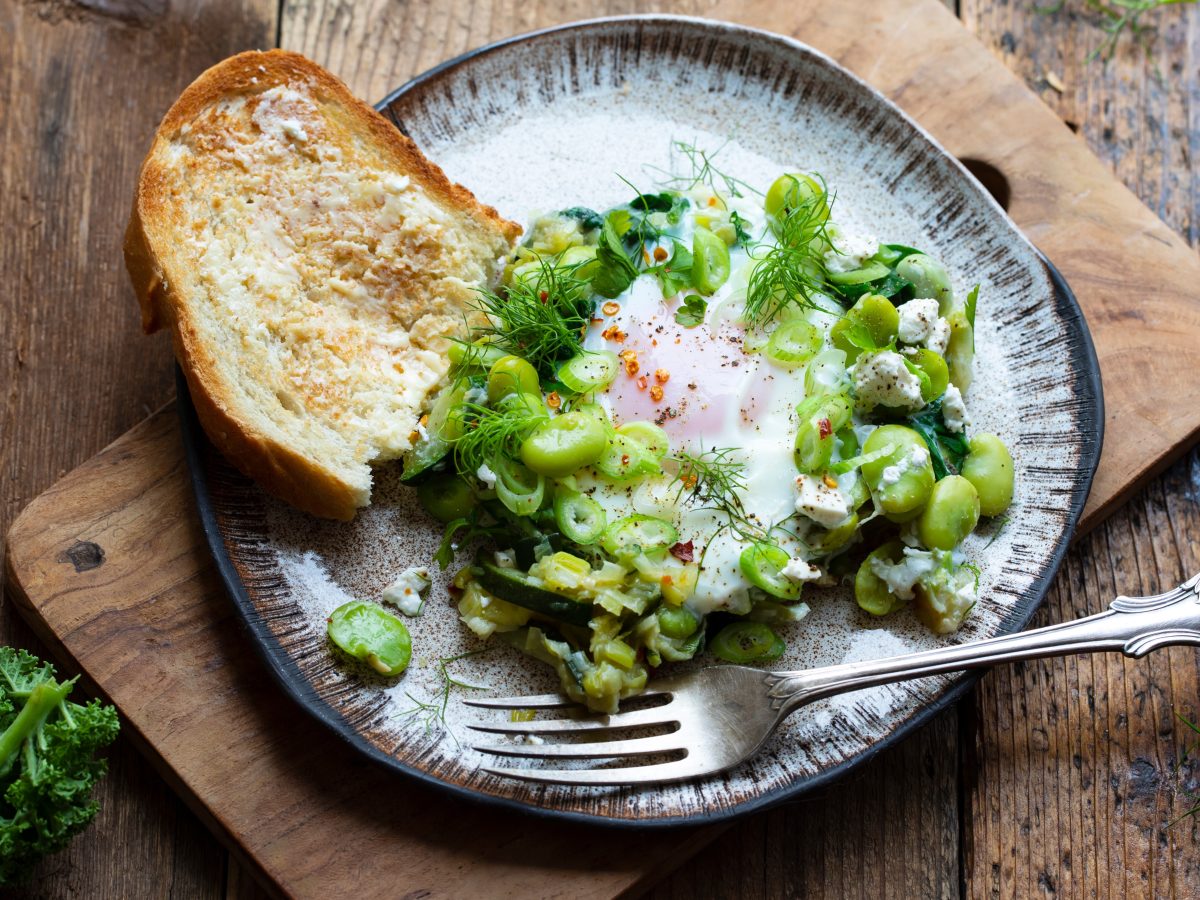 Grünes Shakshuka mit Ackerbohnen, Zucchini, Dill und Spinat auf einem Teller mit gebuttertem Toast.