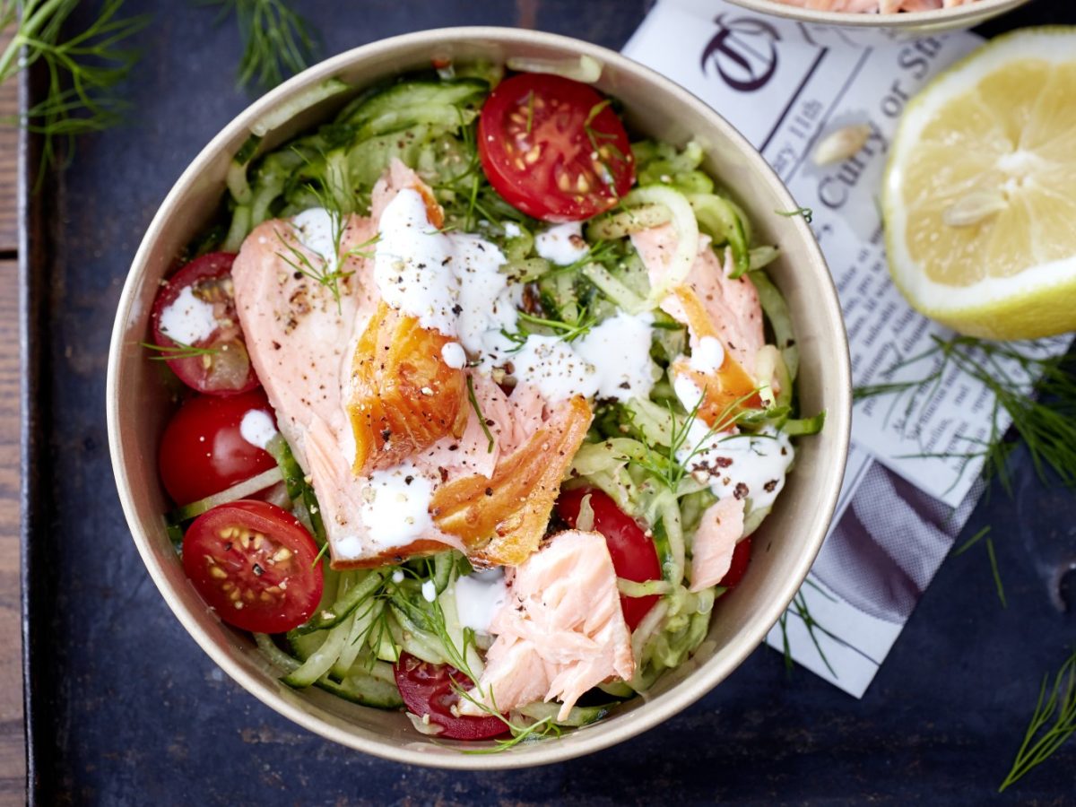 Gurkenspaghetti und Lachs in einem tiefen Teller, daneben eine halbe Zitrone, dunkler Untergrund.