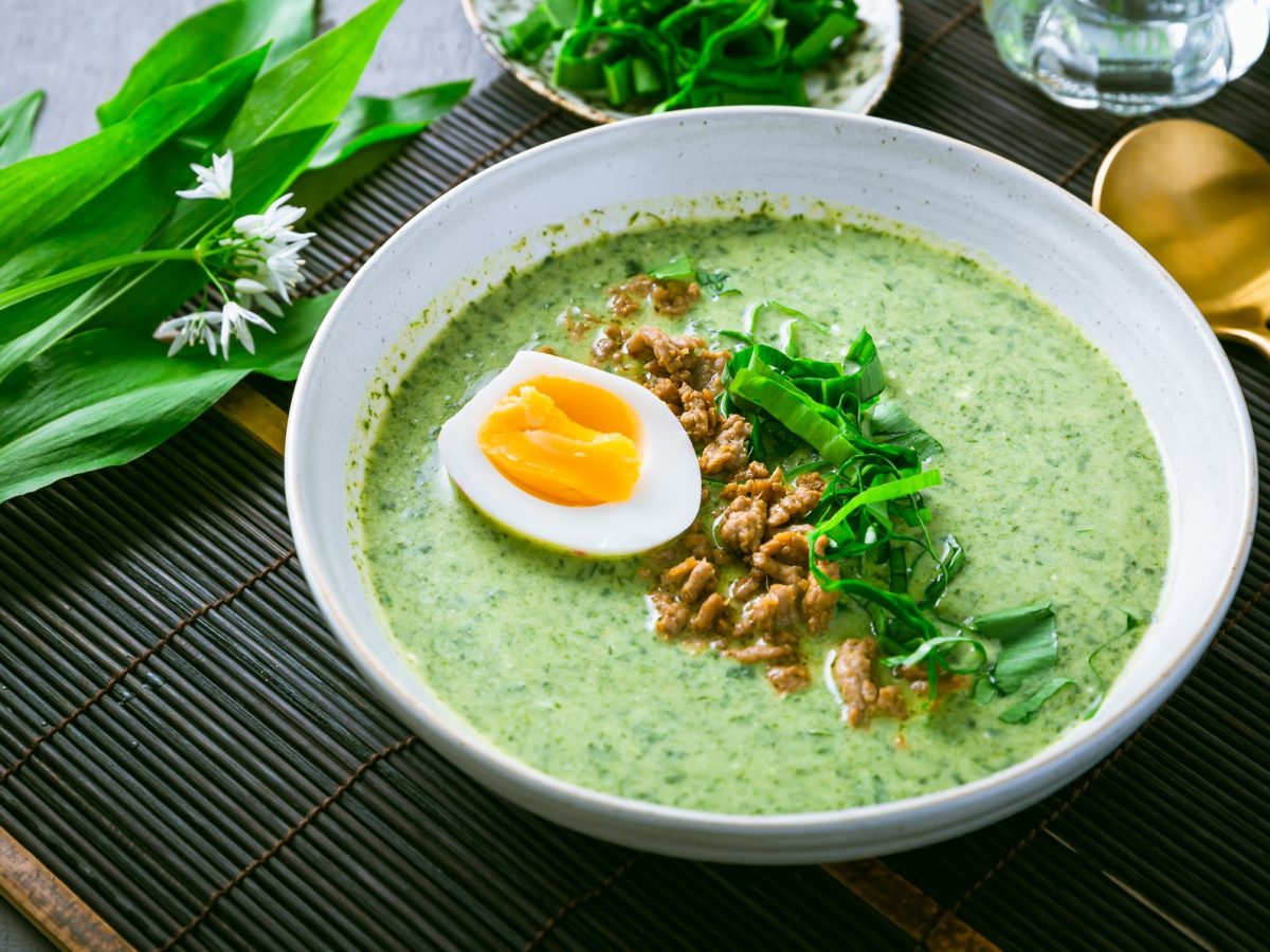 Ein weißer Teller mit der grünen Suppe auf dunklem Untergrund mit frischem Bärlauch und einem goldenen Löffel. Alles von schräg oben fotografiert.