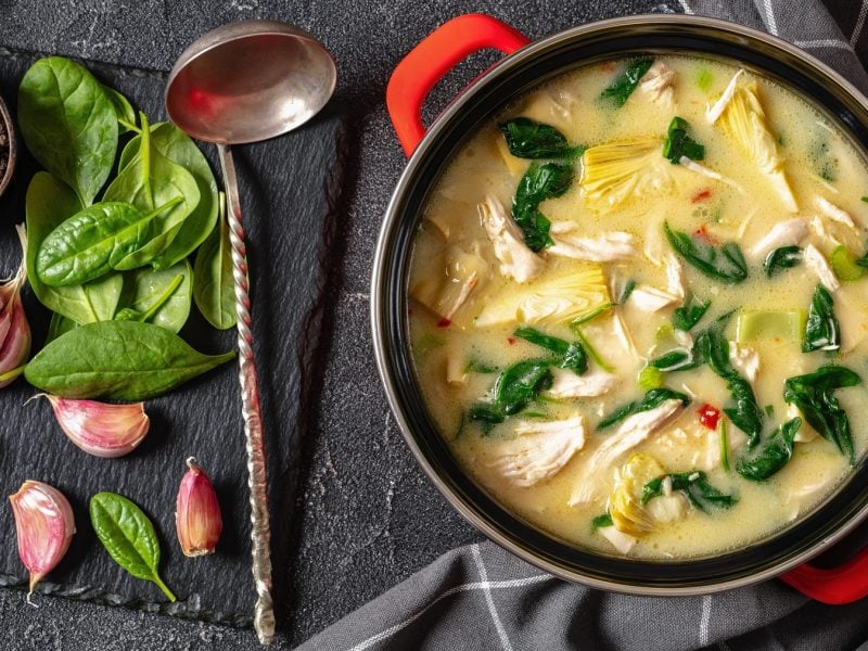 Hähnchensuppe mit Spinat und Artischocken in einem Topf. Daneben Suppenkelle und Zutaten.