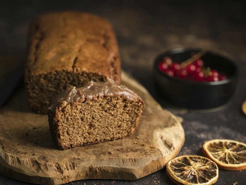 Der Kuchen auf einem Holzbrett und dunklem Untergrund. Daneben getrocknete Orangenscheibe und eine Schale mit roten Beeren.