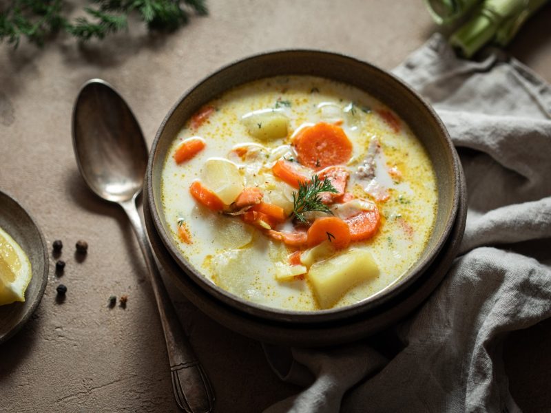 Eine braune Schüssel mit der Lachs-Kartoffel-Suppe auf einem grauen Tisch mit weiteren Gemüsezutaten. Alles von schräg oben fotografiert.