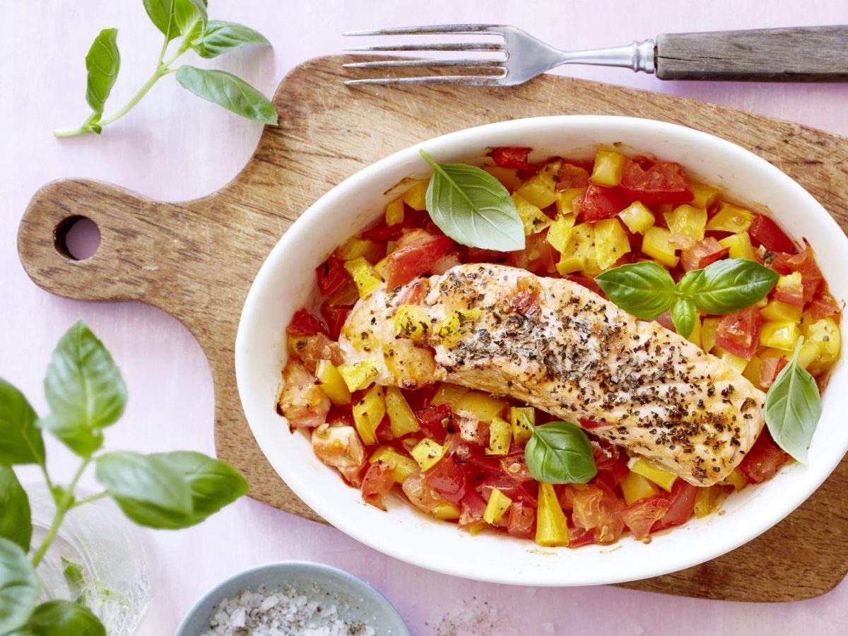 Lachs mit Basilikum und Gemüse in weißer Ofenform auf einem Holzbrett mit hellem Untergrund, daneben Gabel und Basilikumpflanze.