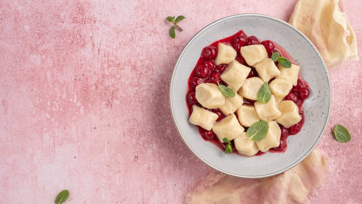 Leniwije vareniki mit Kirschen und Minzblätter auf einem grauen Teller vor rosa Hintergrund.