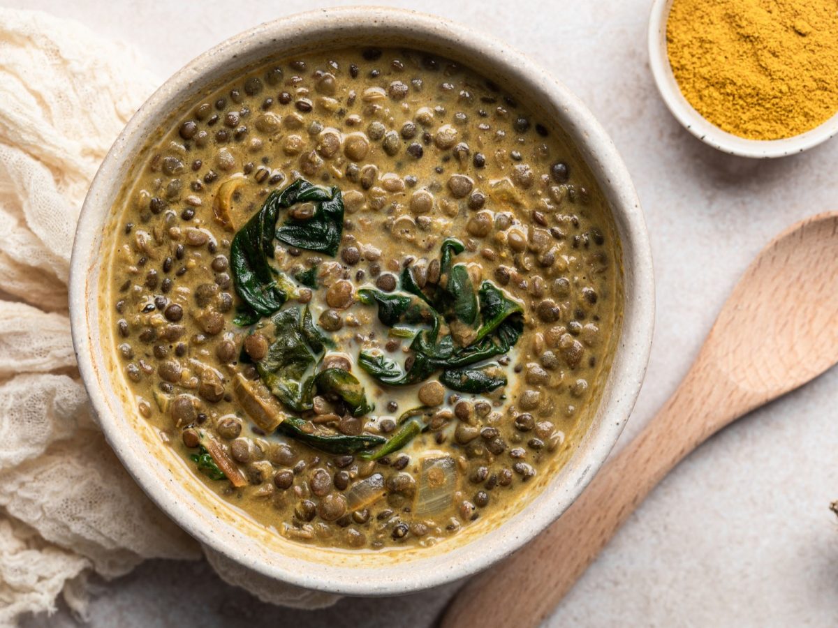 Eine beige Schale mit der Suppe auf einem hellen Tisch, mit Currypulver, Holzkelle und weißem Tuch. Alles direkt von oben fotografiert.