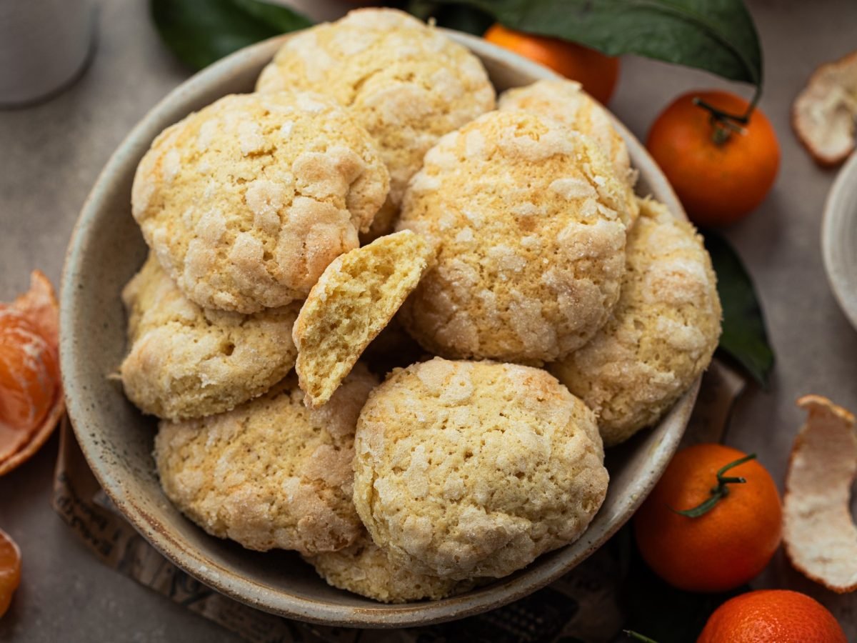 Mehrere Mandarinen-Crinkle-Cookies in einer grauen Schale auf einem grauen Tisch von schräg oben fotografiert. Daneben viele geschälte Mandarinen.