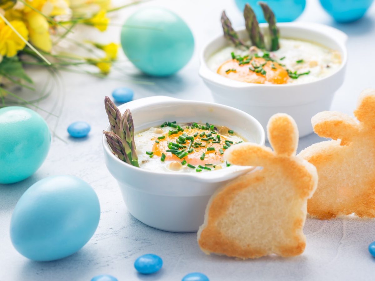 Zwei weiße Schalen mit dem Gericht auf einem hellen Tisch. Daneben viel blaue Osterdeko ein gelber Blumenstrauß und zwei Toastbrote ausgestochen als Osterhase. Alles von der Seite fotografiert.