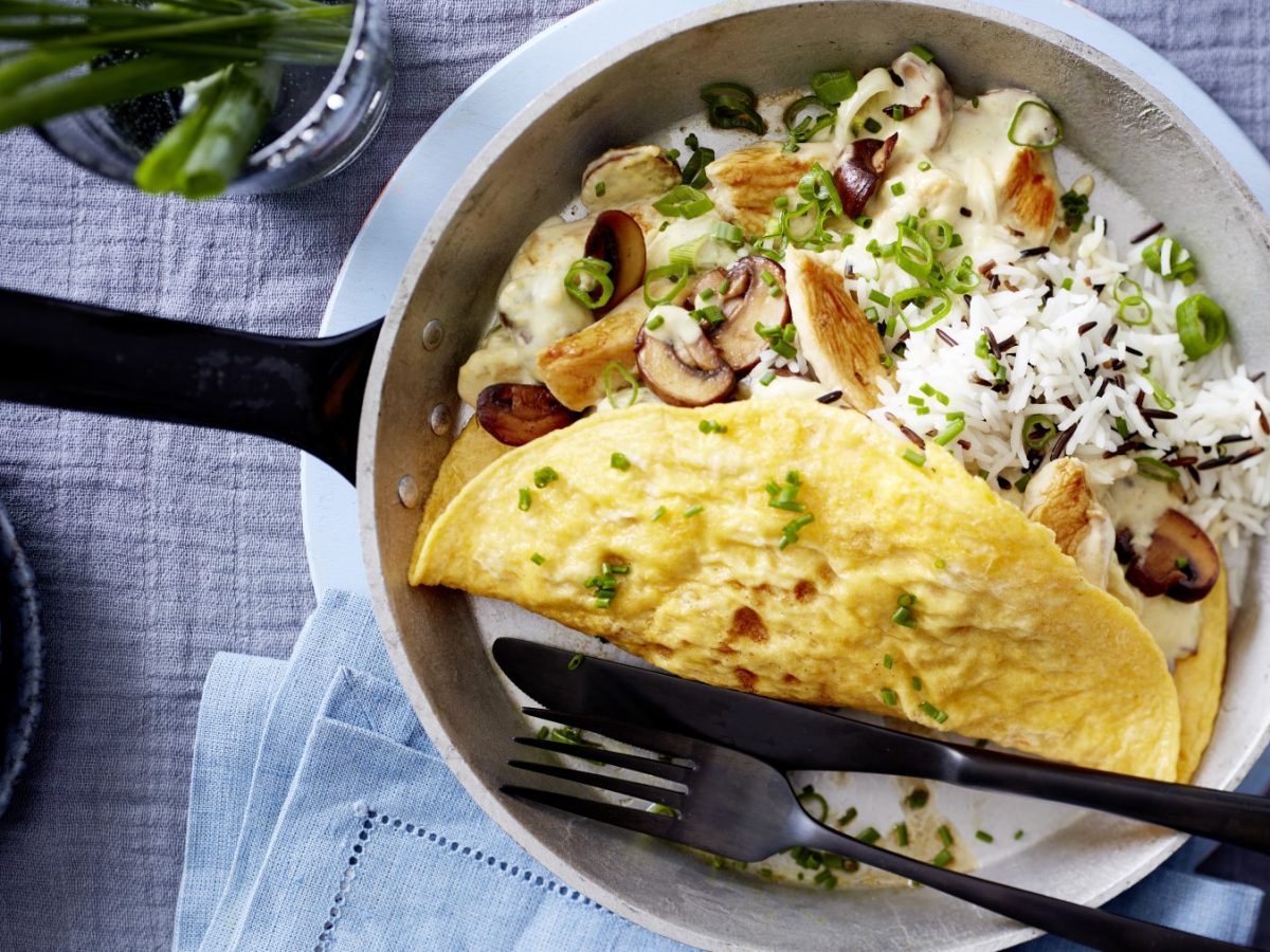 Omelett mit Hähnchen-Pilz-Ragout in einer Pfanne auf einem grauen Hintergrund. Oben auf dem Foto Schnittlauch, unten auf dem Foto ein Geschirrtuch und links ein kleiner Teller.