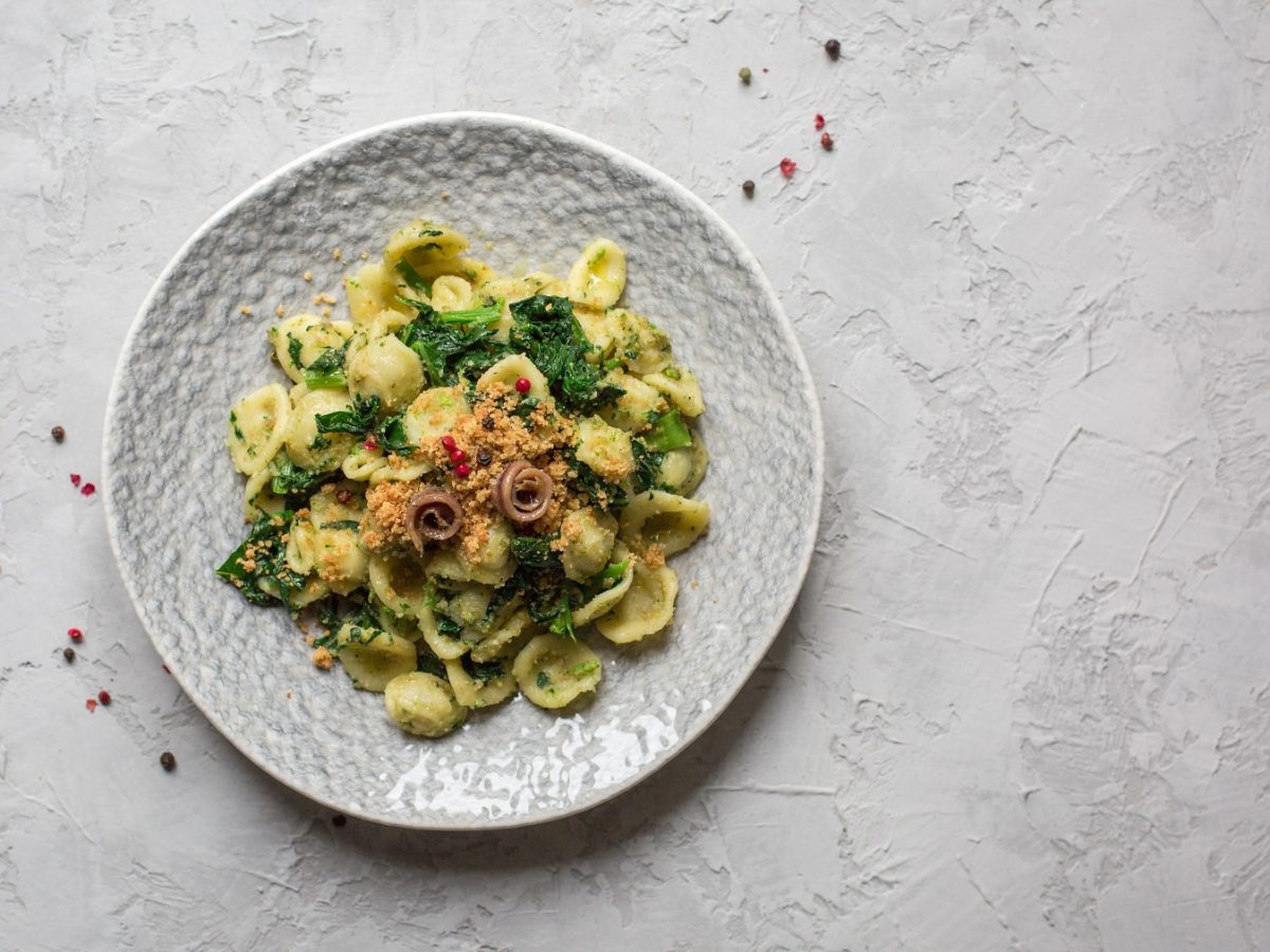 Draufsicht von Orecchiette mit Brokkoli und Sardellen auf einem weißen Teller. Daneben rosa Pfefferkörner.