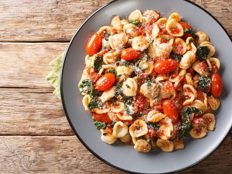 Draufsicht auf Orecchiette mit Spinat und Tomaten auf einem grauen Teller auf einem Holztisch.