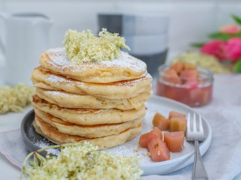 Gestapelte Pancakes und daneben das Rhabarber-Holunderblüten-Kompott. Deko aus frischen Holunderblüten.