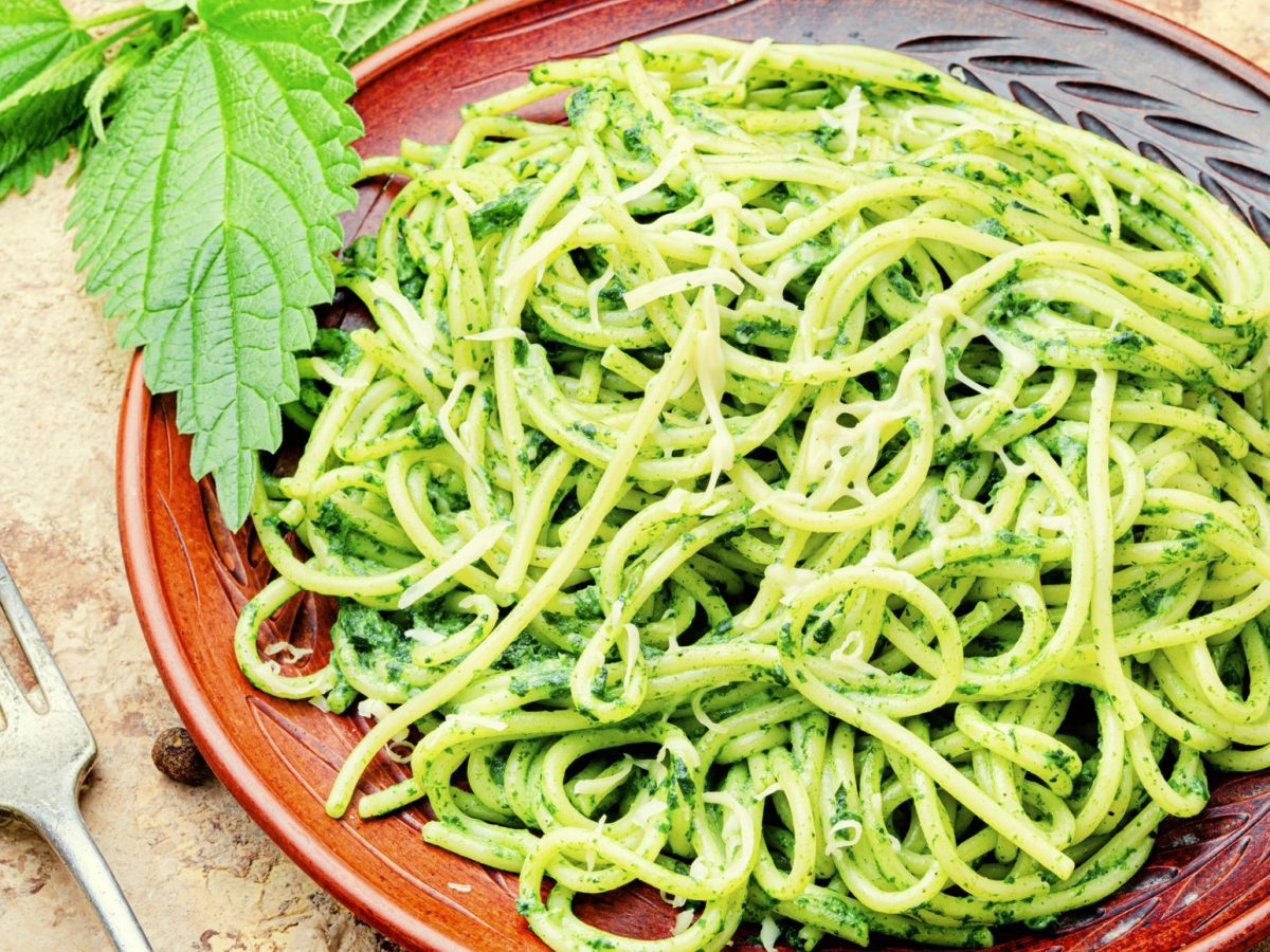 Ein brauner Teller mit dem Brennnesselpesto auf einem Holztisch mit frischer Brennnessel und Parmesan.