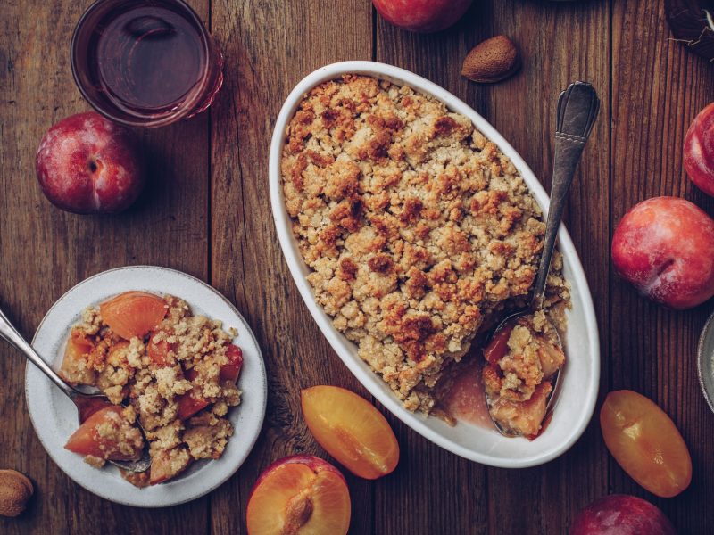 Eine weiße Auflaufform mit dem Pflaumen-Crumble auf einem rustikalen Holztisch mit angeschnittenen Pflaumen. Alles direkt von oben fotografiert.