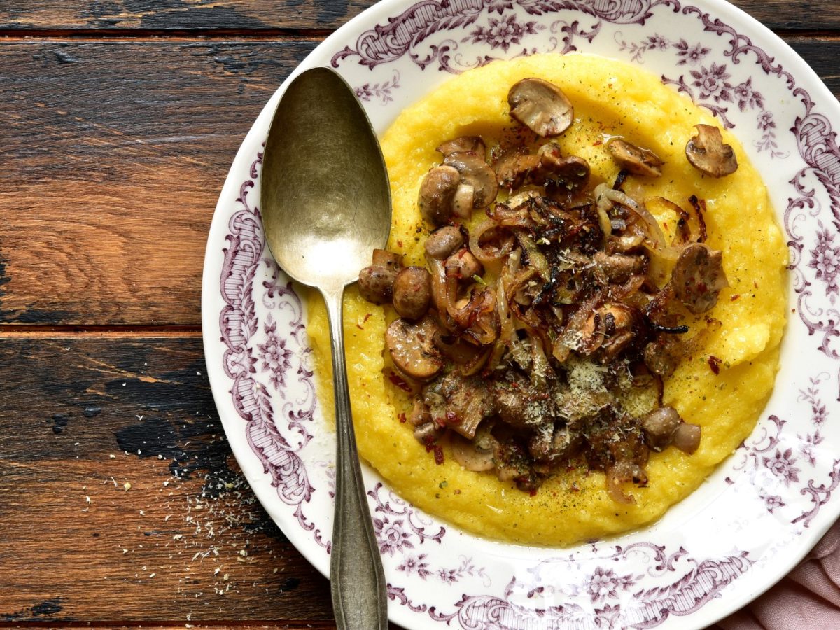 Polenta mit karamellisierten Pilzen in einem Teller. Links ein Löffel. Der Hintergrund ist hölzern.