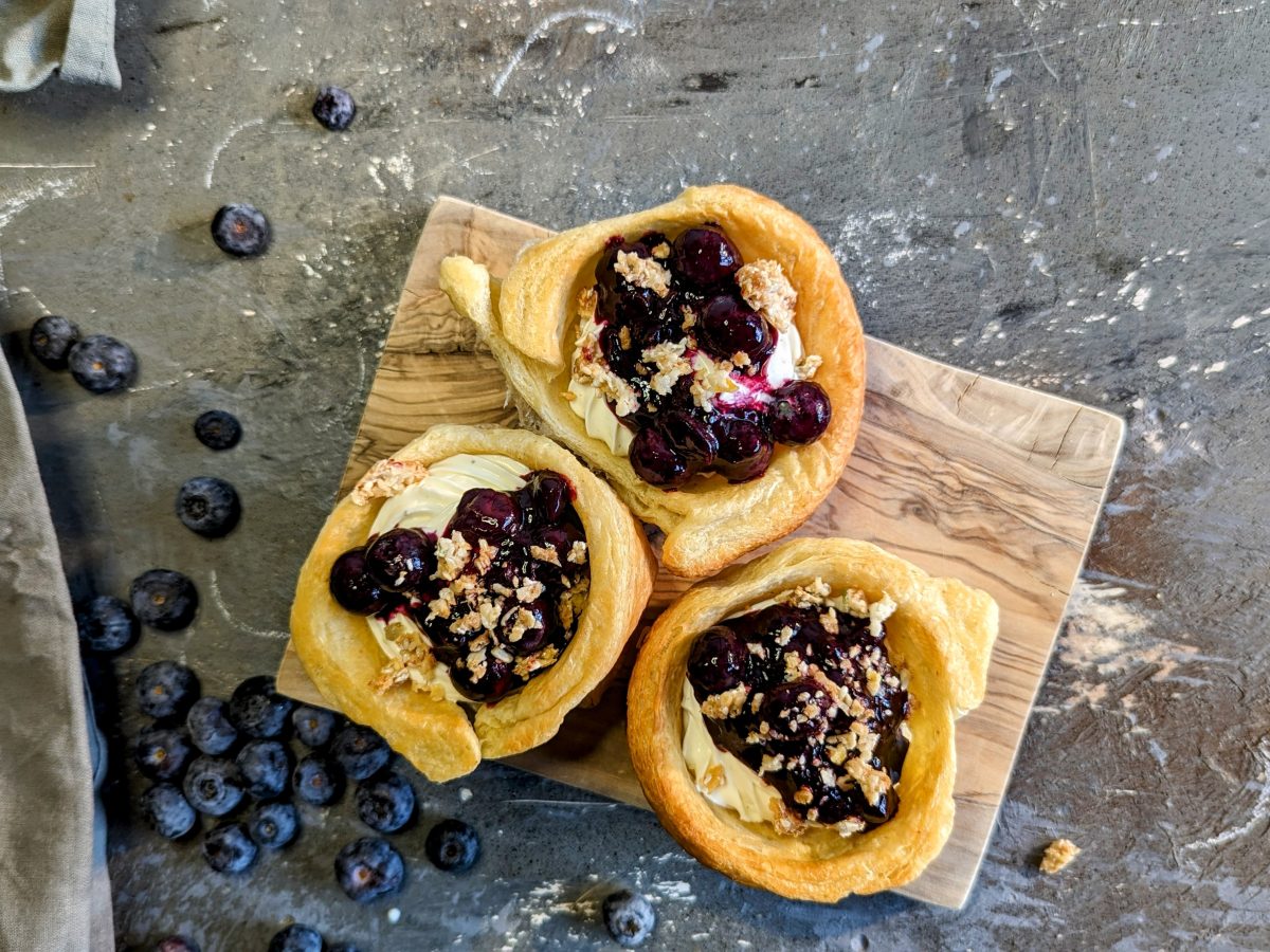 Drei gefülkte Cruffins mit Blaubeerragout auf einem kleinen Holzbrett und einem Steintisch mit Blaubeeren drumherum. Alles direkt von oben fotografiert.