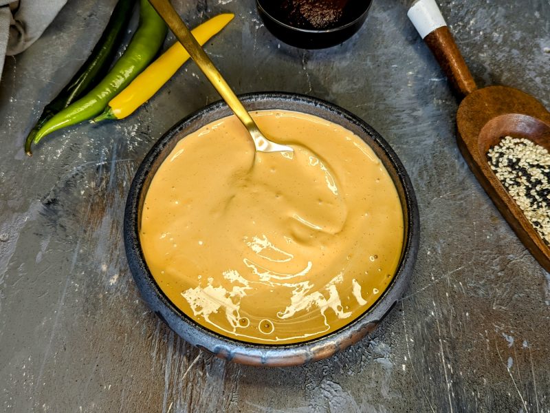 Eine graue Schale mit der koreanischen Chili-Mayonnaise auf einem Steintisch mit Sesam und Chilischoten. Alles von oben fotografiert.