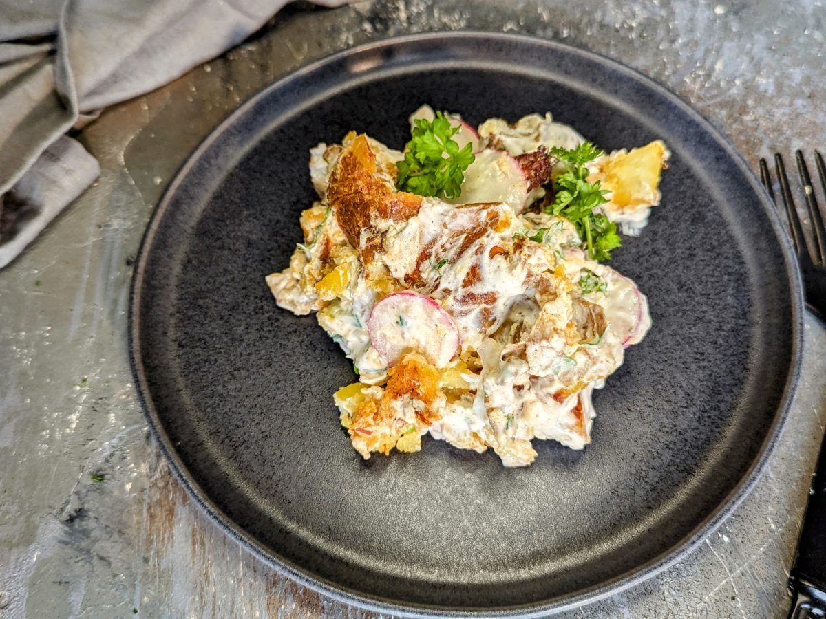 Ein schwarzer Teller mit dem Smashed Potato Salad und schwarzem Besteck sowie grauem Tuch auf einem Steintisch. Alles von schräg oben fotografiert.