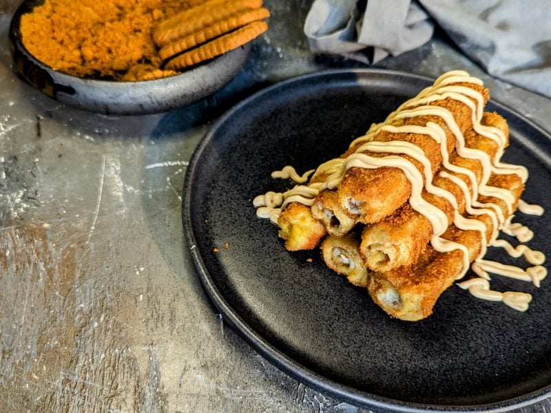 Ein schwarzer Teller mit den Biscoff-French-Toast-Rolls auf einem grauen Tisch mit den Keksen im Hintergrund.