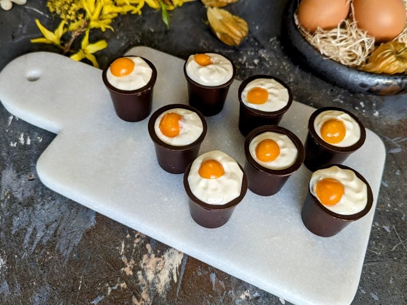 Mehrere Schokobecher mit der Eierlikör-Creme auf einem kleinen weißen Marmorbrett und grauem Tisch. Im Hintergrund Osterdekoration und alles von schräg oben fotografiert.