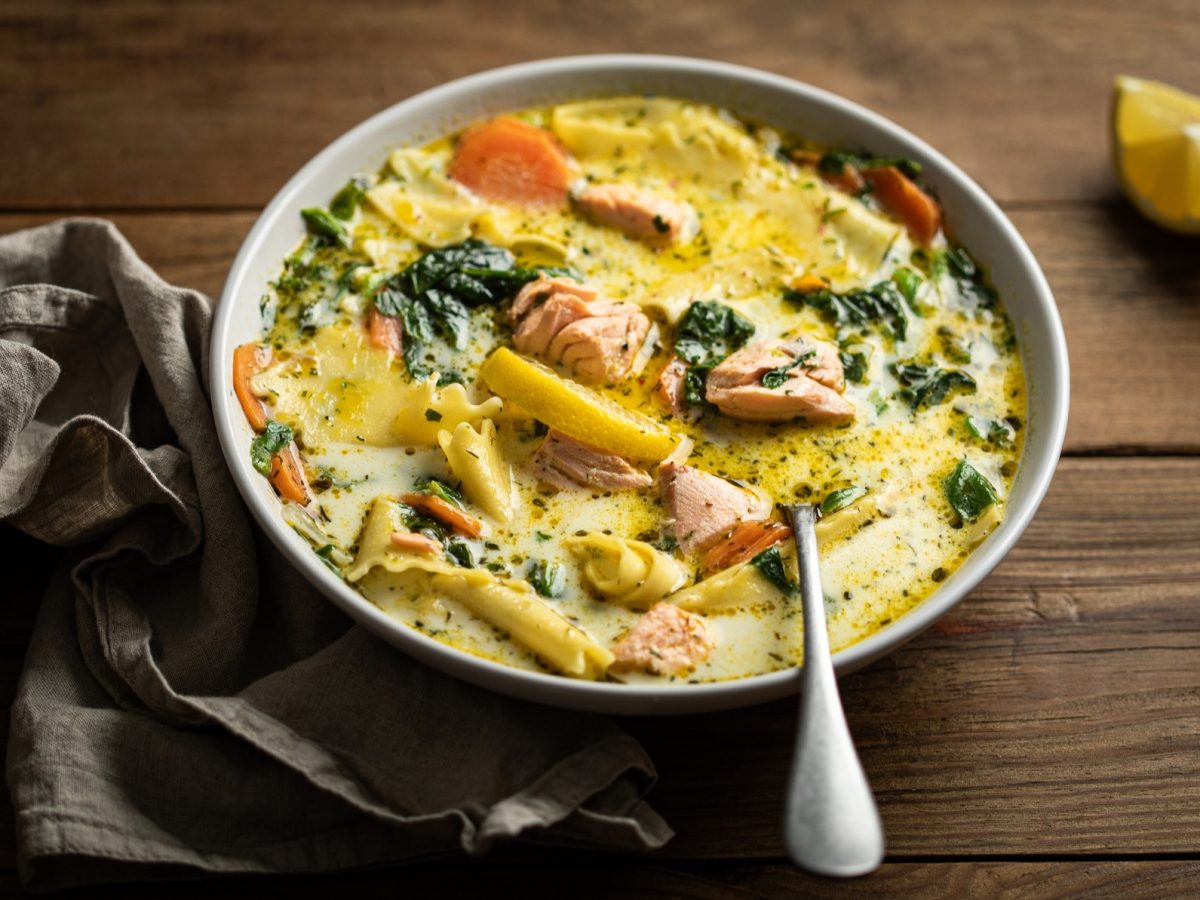 Eine helle Schale mit der Suppe auf einem rustikalen Holztisch mit grauem Tuch und Zitronenecke. Alles von schräg oben fotografiert.