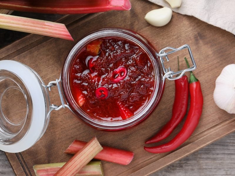 Rhabarber-Chutney in einem Glas. Daneben frische Chilischoten, Knoblauch und Rhabarberstangen.