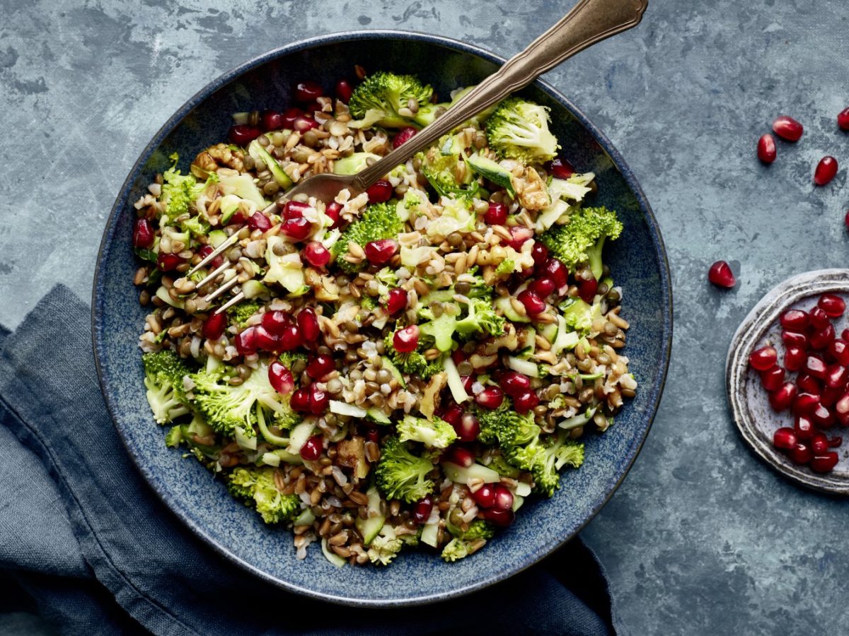 Linsen-Brokkoli-Bowl in einer schwarzen Schüssel auf grauem Hintergrund. Rechts eine kleine Schüssel mit Granatapfelkernen und links unten ein graues Geschirrtuch.