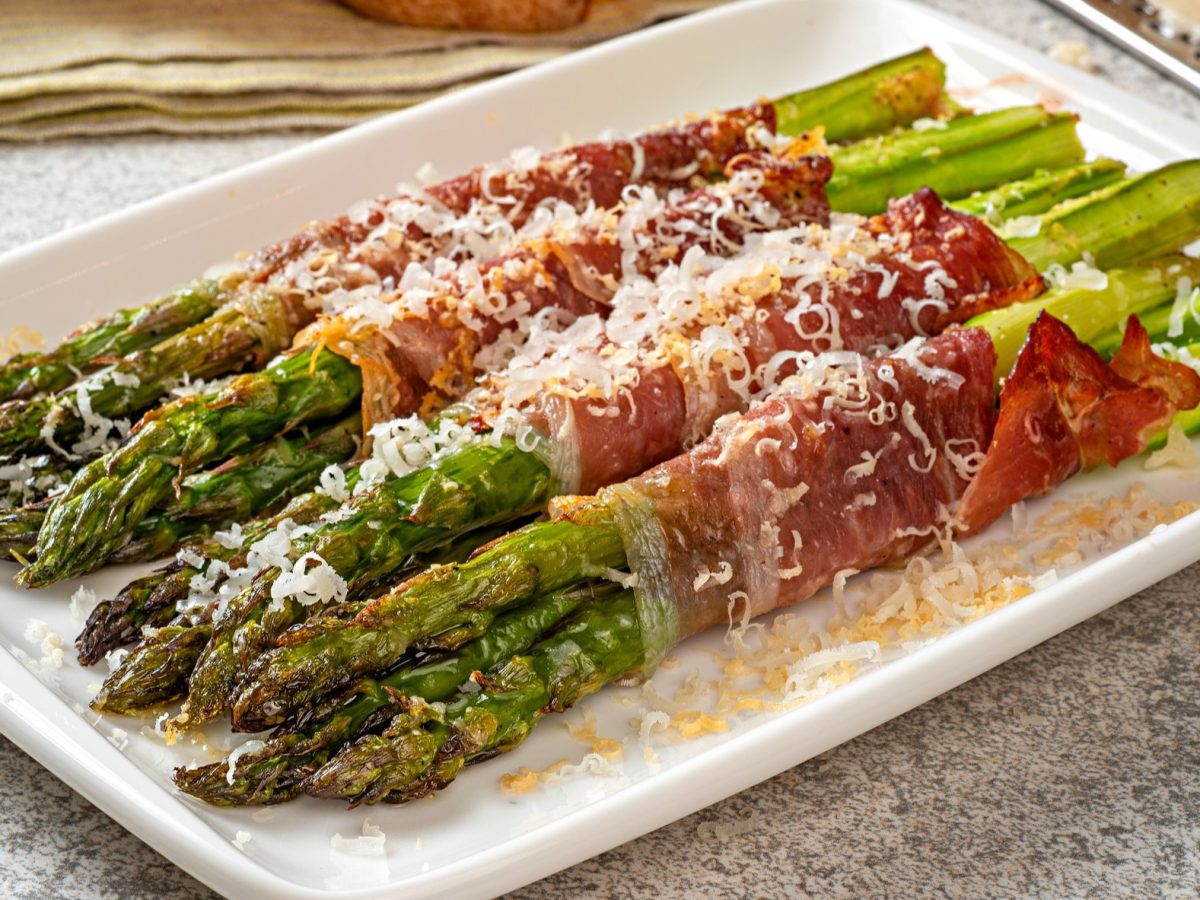 Ein weißer, viereckiger Teller mit den Spargel-Schinken-Päckchen und gehobeltem Parmesan in der Nahaufnahme auf grauen Untergrund. Scharfer Fokus auf die Spargelköpfe.