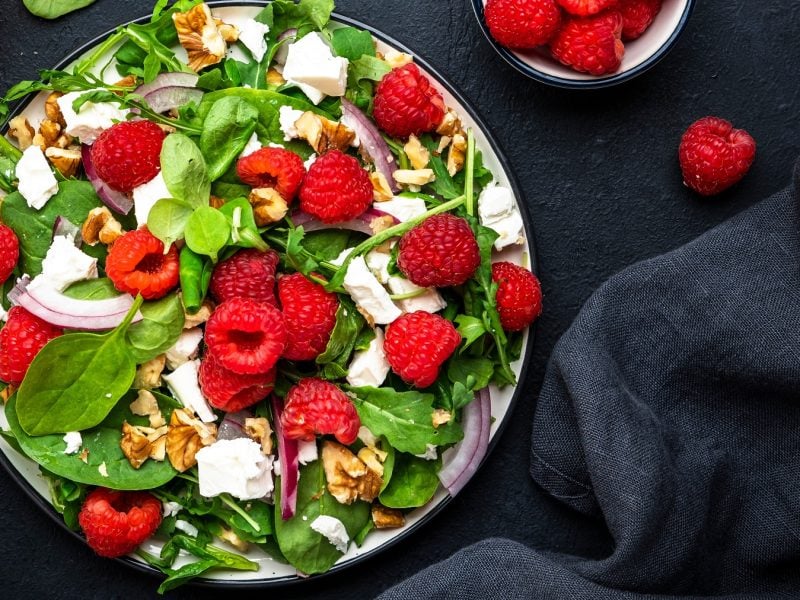 Spinat-Salat mit Himbeeren, Ziegenkäse, Walnüssen und roten Zwiebeln auf einem Teller.