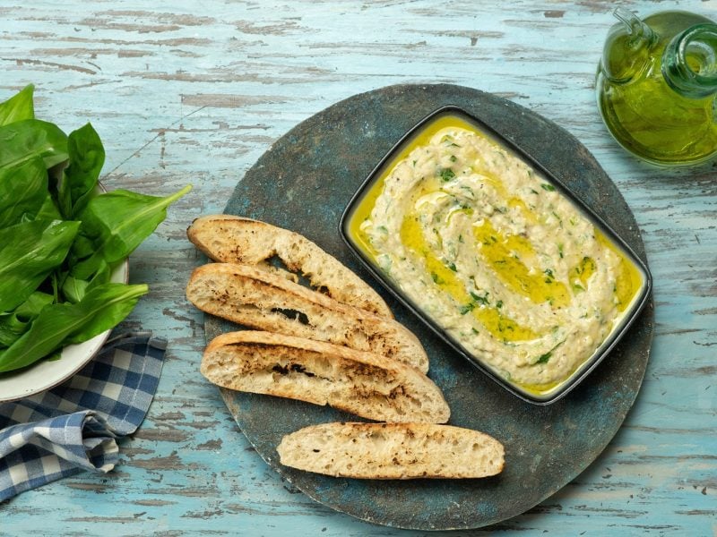 Draufsicht auf Zucchini-Dip auf einem teller mit Brot. Daneben frischer Bärlauch und Olivenöl in einer Karaffe.