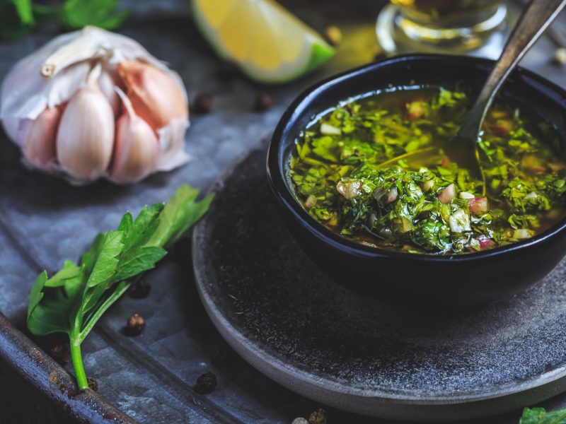 Ein schwarzes Schälchen mit dem Bärlauch-Chimichurri auf dunkelblauem Untergrund, mit Kräutern und Knoblauch. Alles von schräg oben und recht nahe fotografiert.