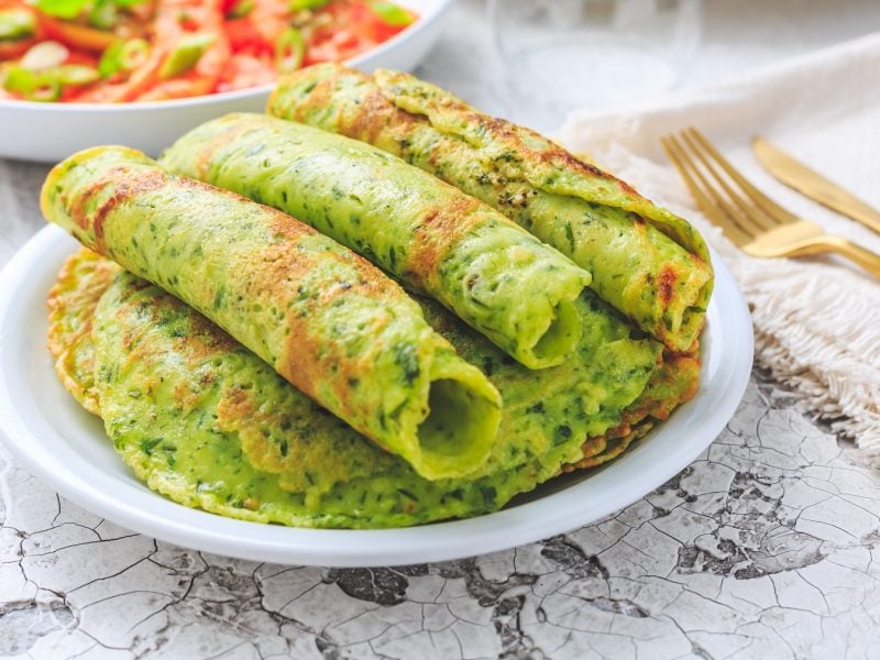 Mehrere Bärlauch-Pfannkuchenröllchen auf einem weißen Teller und hellem Marmoruntergrund. Im Hintergrund eine weiße Schale mit Tomatensalat erkennbar.