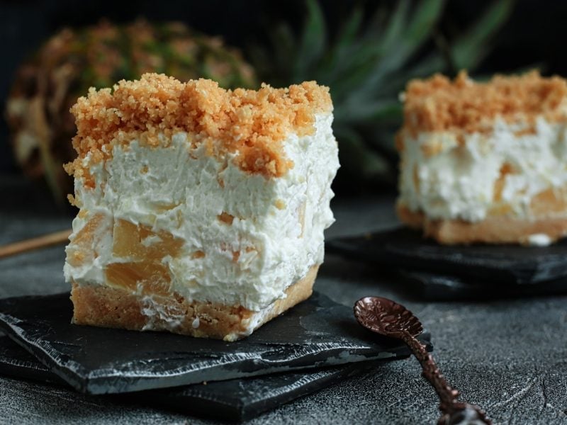 Zwei Stücke mit dem Kuchen auf schwarzen Schieferplatten und dunklem Tisch. Im Hintergrund unscharf eine Ananas und dunkler Hintergrund zu erkennen.