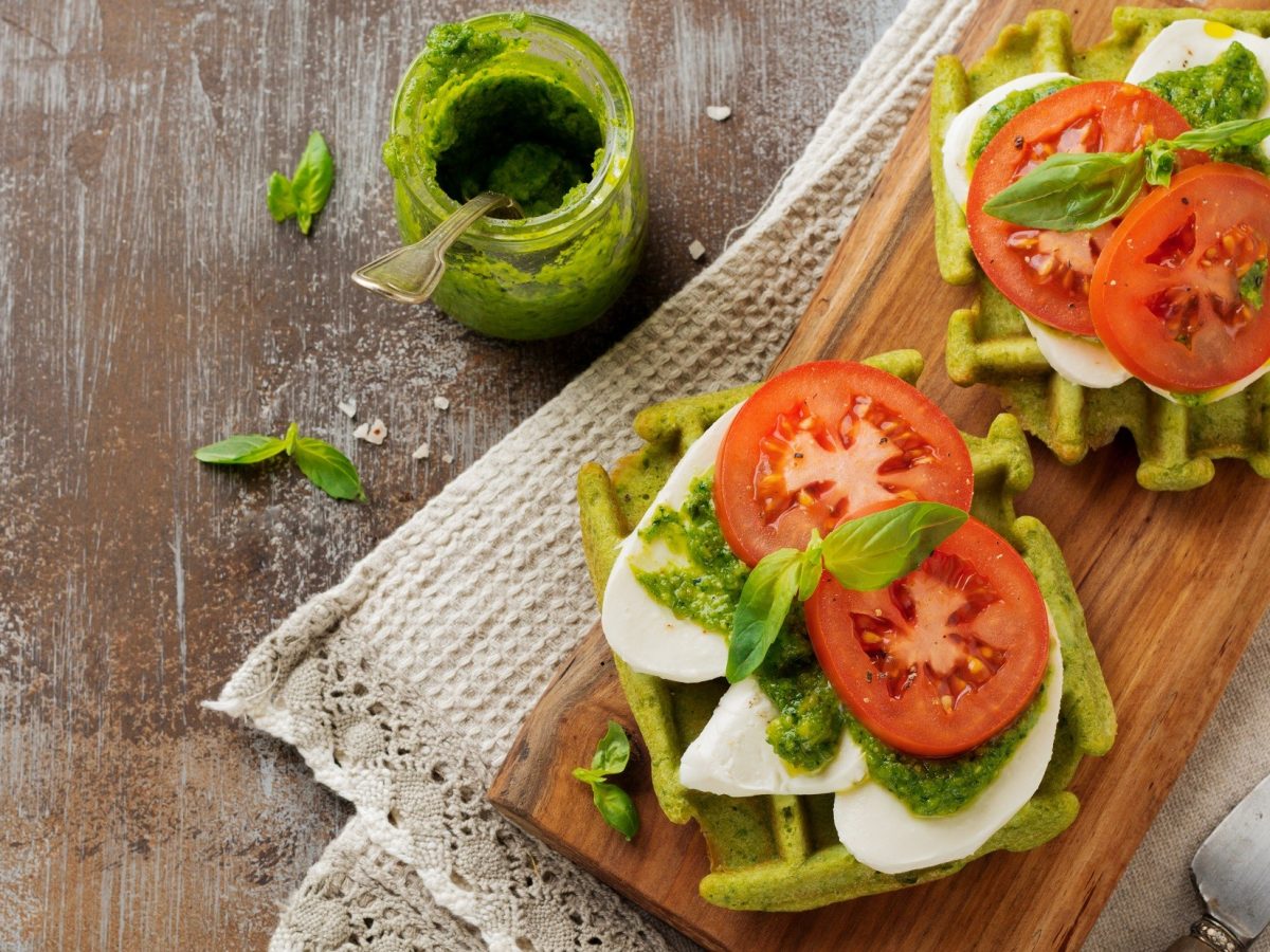 Draufblich: Ein Holzbrettchen mit zwei Caprese-Waffeln, die mit Mozzarella, Basilikum, Pesto und frische Tomaten belegt sind.