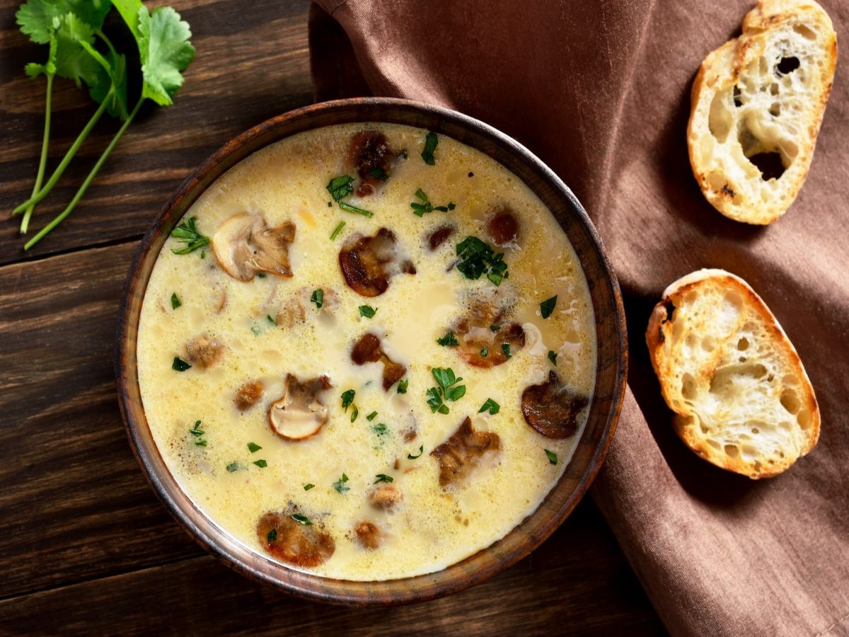 Draufsicht: Eine Schüssel Champignon-Suppe mit Gruyère steht auf einem dunklen, rustikalen Untergrund aus Holz. Daneben liegen zwei geröstete Weißbrotscheiben.