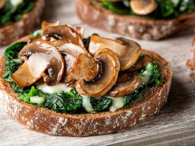 Draufsicht, schräg: ein Cheese Sandwich mit Spinat und Champignons aus der Pfanne liegt auf einem hellen Holzbrett. Im Hintergrund sind drei weitere Brote teilweise zu sehen.