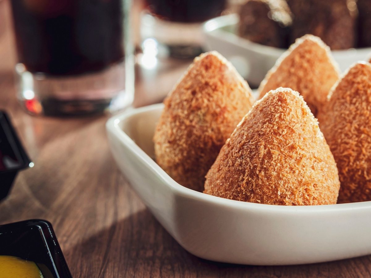 Frontal: Fümf Coxinha de Frango stehen in einer weißen , flachen Schale auf einem Tisch. Es handelt sich um brasilianische, frittierte Snacks aus Hühnchenfleisch.