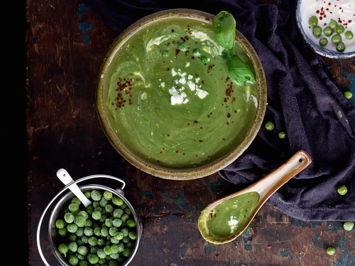 Draufsicht. Eine runde Schüssel Erbsensuppe mit Feta auf einem dunklem Untergrund. Daneben sind eine kleine Schale mit TK-Erbsen, ein Löffel und eine Schale mit einer weißen Creme.