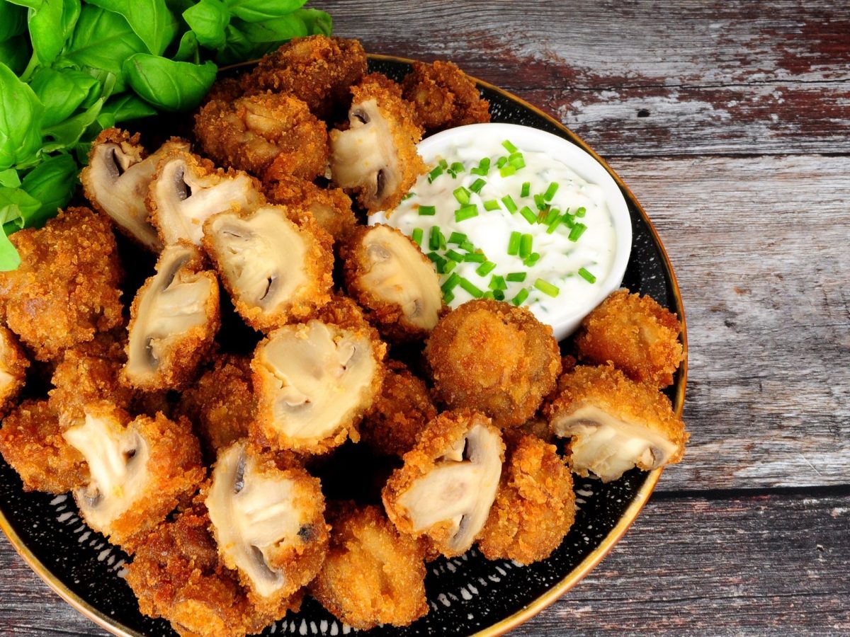 Draufsicht: Eine Teller frittierte Champignons mit einem schälchen weißer Sauce. An einem Rand sind frische Basilikum-Blätter.