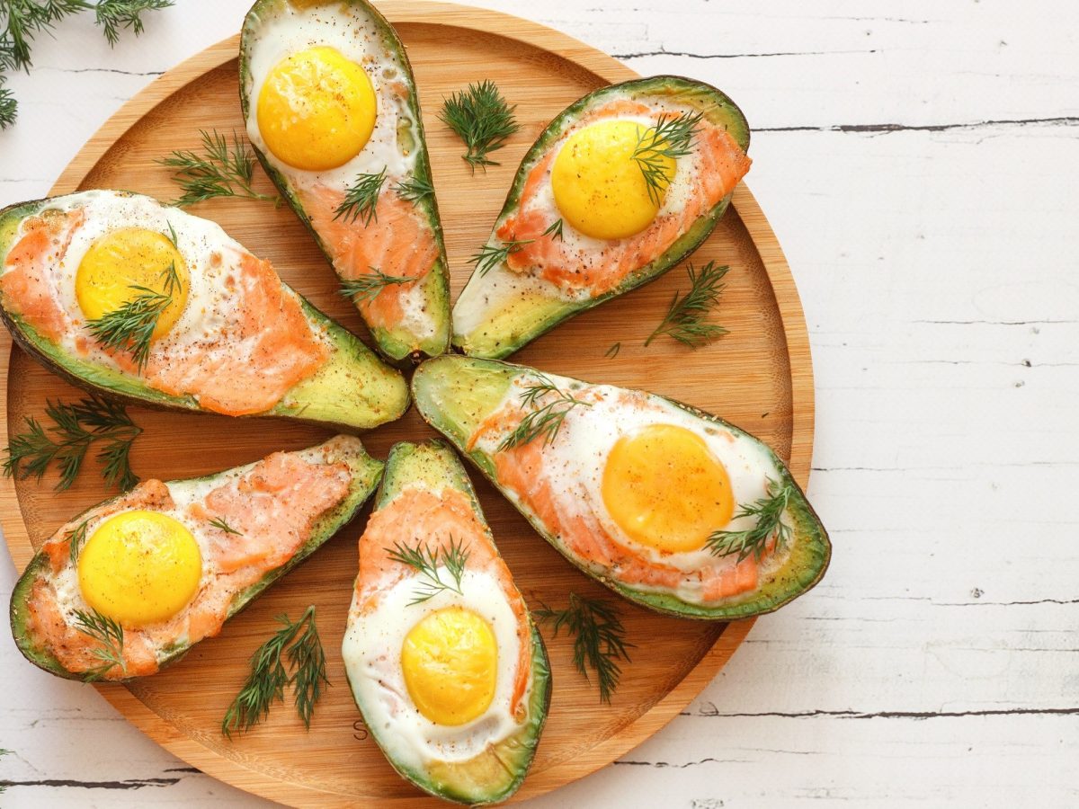 Draufsicht: Sechs gefüllte Avocado mit Lachs und Ei auf einem hellbraunen Teller. Der liegt auf einem weißen Untergrund aus Holz. Drum herum liegen frische Dillspitzen.