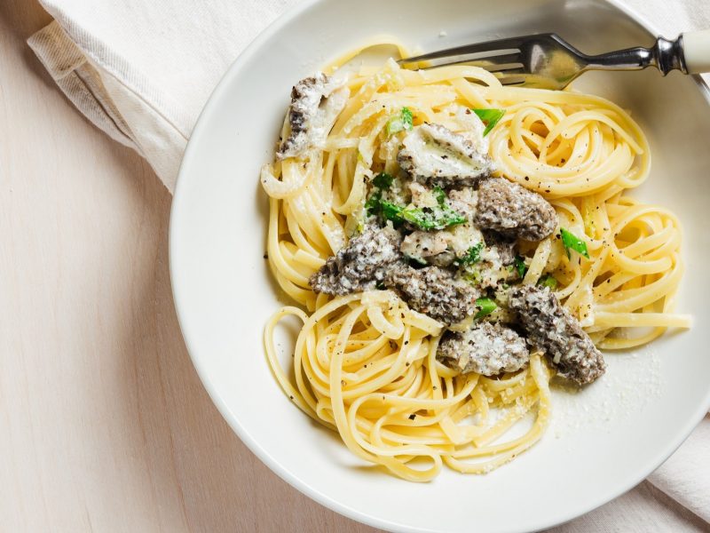 Draufsicht: Ein Teller Linguine mit Morcheln auf einem hellen Untergrund. Daneben liegen ein helles Tuch und eine Gabel.